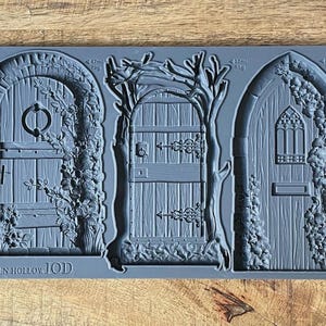 May include: A grey silicone mould depicting three detailed fairy doors. The doors are arched and rectangular, with intricate details like hinges, handles, and surrounding foliage. The text "HIDDEN HOLLOW IOD" is at the bottom.
