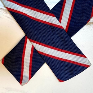 Peut inclure: Cravate bleu marine avec des rayures diagonales rouges et blanches. La cravate est en tissu côtelé et est pliée sur elle-même. Un accessoire classique.