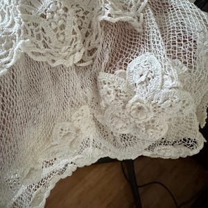 May include: Close-up of a white, intricately crocheted textile. The delicate openwork design features floral and geometric patterns, with a scalloped edge. The texture is soft and airy, suggesting a handmade shawl or decorative piece.