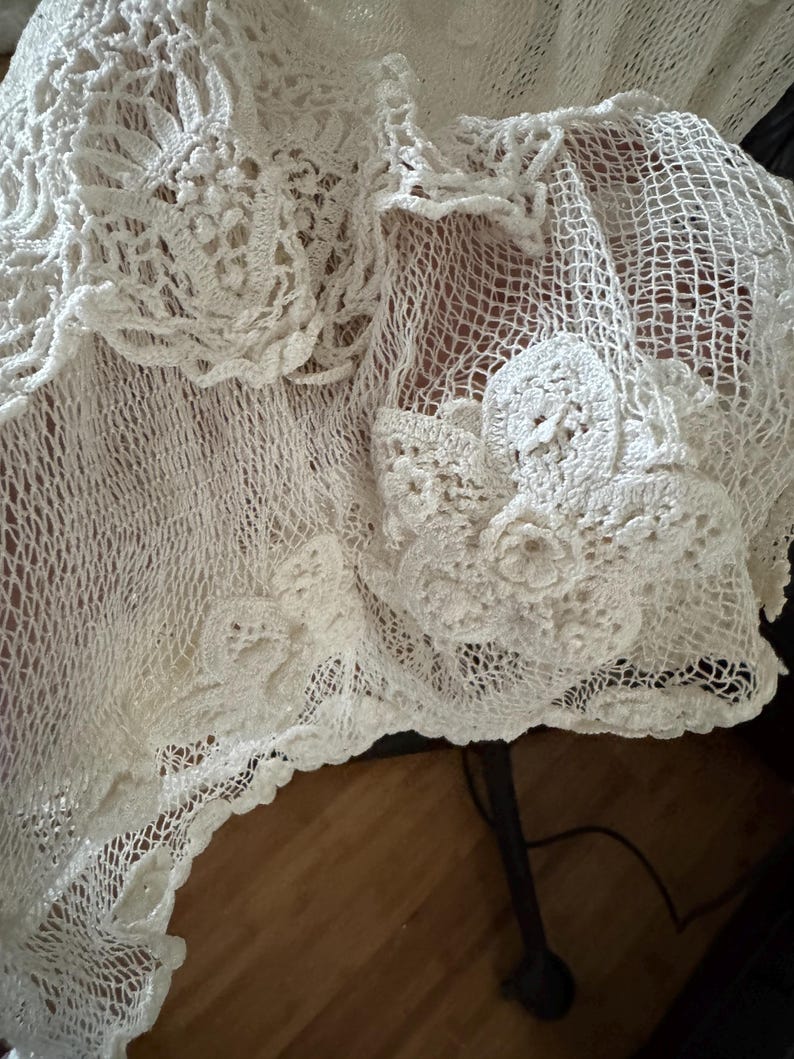 May include: Close-up of a white, intricately crocheted textile. The delicate openwork design features floral and geometric patterns, with a scalloped edge. The texture is soft and airy, suggesting a handmade shawl or decorative piece.