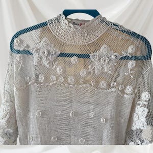 May include: A white lace top with a high, crochet collar and long sleeves. The top is adorned with floral crochet appliques across the chest and sleeves. The delicate, open-weave fabric is visible throughout the garment.