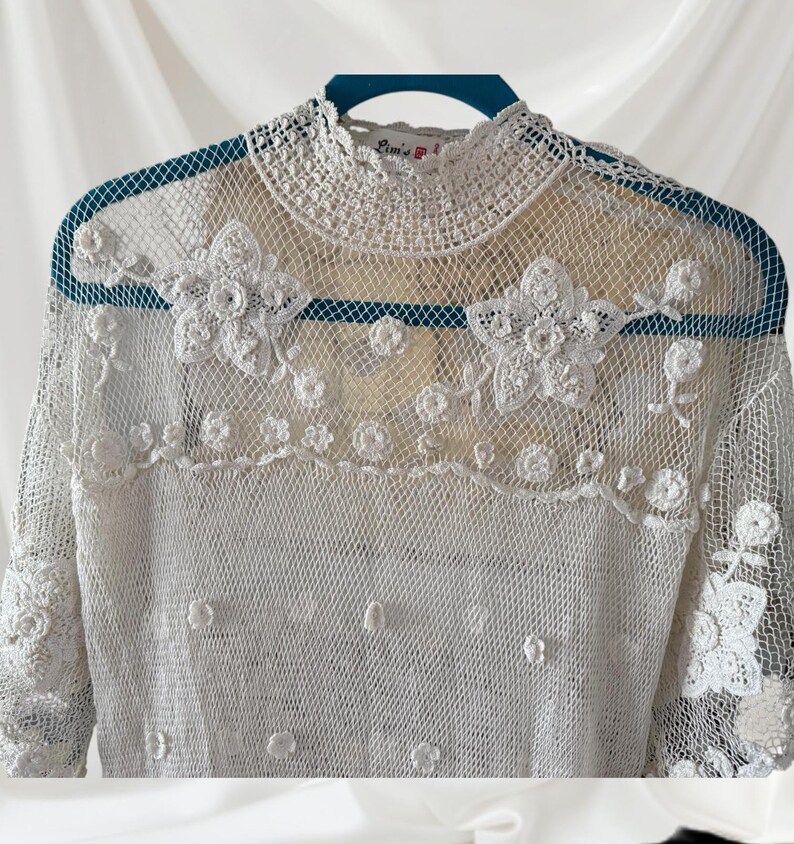 May include: A white lace top with a high, crochet collar and long sleeves. The top is adorned with floral crochet appliques across the chest and sleeves. The delicate, open-weave fabric is visible throughout the garment.