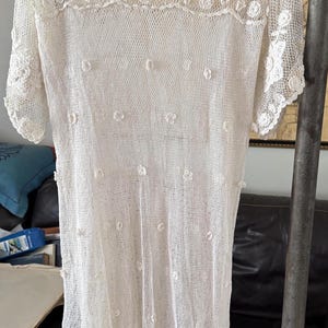 May include: A white, open-weave dress with a floral lace yoke and sleeves. The dress features small, raised floral accents and a scalloped lace hem. The dress is hanging on a blue hanger.