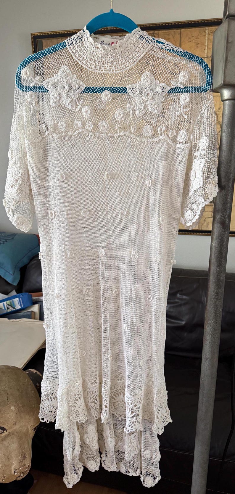 May include: A white, open-weave dress with a floral lace yoke and sleeves. The dress features small, raised floral accents and a scalloped lace hem. The dress is hanging on a blue hanger.