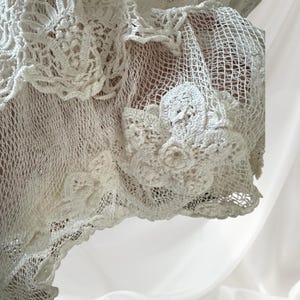 May include: Close-up of a white, open-weave crochet garment. The garment features intricate floral and geometric patterns, with a delicate, lacy texture. The crochet work is detailed and appears handmade, showcasing a classic, vintage style.