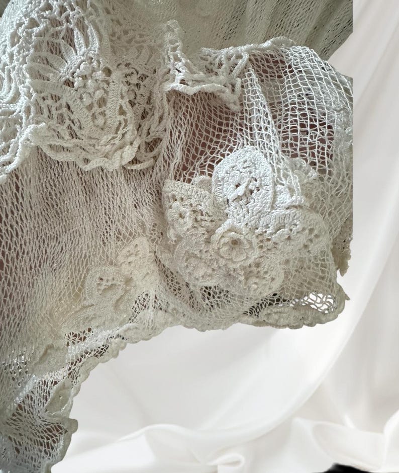 May include: Close-up of a white, open-weave crochet garment. The garment features intricate floral and geometric patterns, with a delicate, lacy texture. The crochet work is detailed and appears handmade, showcasing a classic, vintage style.