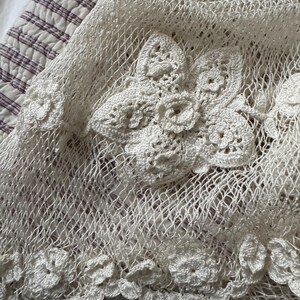 May include: A close-up view of a cream-coloured crocheted textile. The textile has a net-like pattern with large, three-dimensional floral embellishments. The flowers are intricately detailed, adding texture. The edges are also adorned with smaller floral accents.