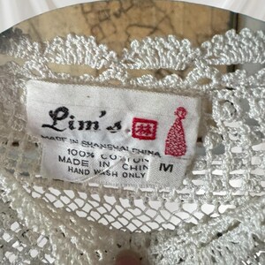 May include: A close-up of a white, crocheted garment with a label reading "Lim's" and "Made in Shanghai China." The label also states the garment is 100% cotton and size M. The garment features a delicate, openwork design.