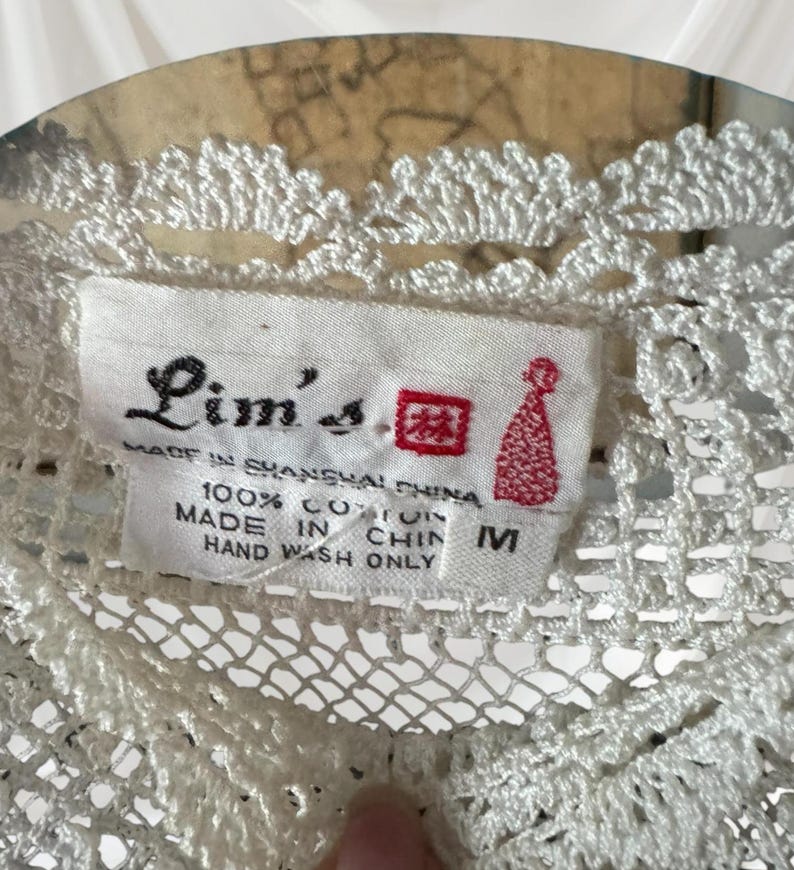 May include: A close-up of a white, crocheted garment with a label reading "Lim's" and "Made in Shanghai China." The label also states the garment is 100% cotton and size M. The garment features a delicate, openwork design.