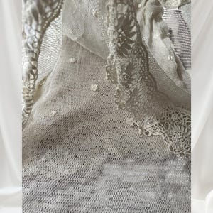 May include: Close-up of a cream-coloured lace fabric with intricate floral and geometric patterns. The delicate material is layered, showcasing a variety of textures and designs. The edges are finished with scalloped lace trim, adding a touch of elegance.