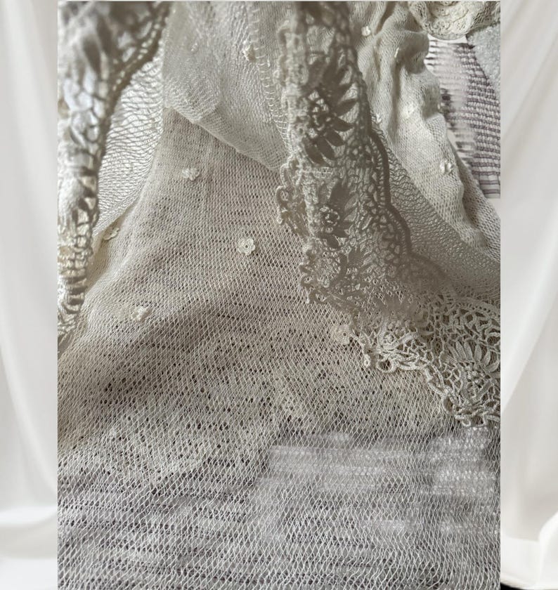 May include: Close-up of a cream-coloured lace fabric with intricate floral and geometric patterns. The delicate material is layered, showcasing a variety of textures and designs. The edges are finished with scalloped lace trim, adding a touch of elegance.