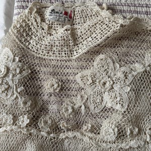 May include: A cream-coloured, open-weave crochet top with floral and circular detailing. The top features a high, rounded neckline and a delicate, lacy texture. The label reads "Lim's".