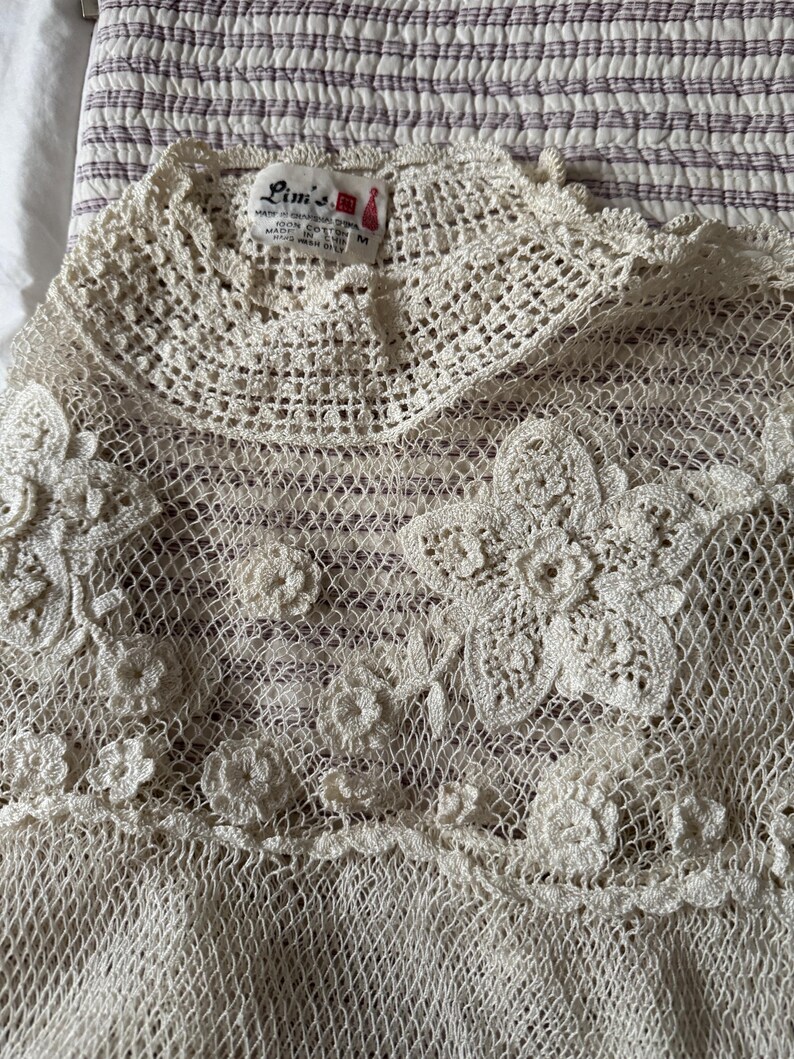 May include: A cream-coloured, open-weave crochet top with floral and circular detailing. The top features a high, rounded neckline and a delicate, lacy texture. The label reads "Lim's".
