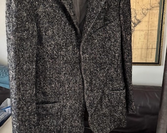 May include: A tweed blazer in shades of gray and black hangs on a blue hanger. The jacket has a two-button closure, notched lapels, and two front pockets. The interior lining is a dark gray color.