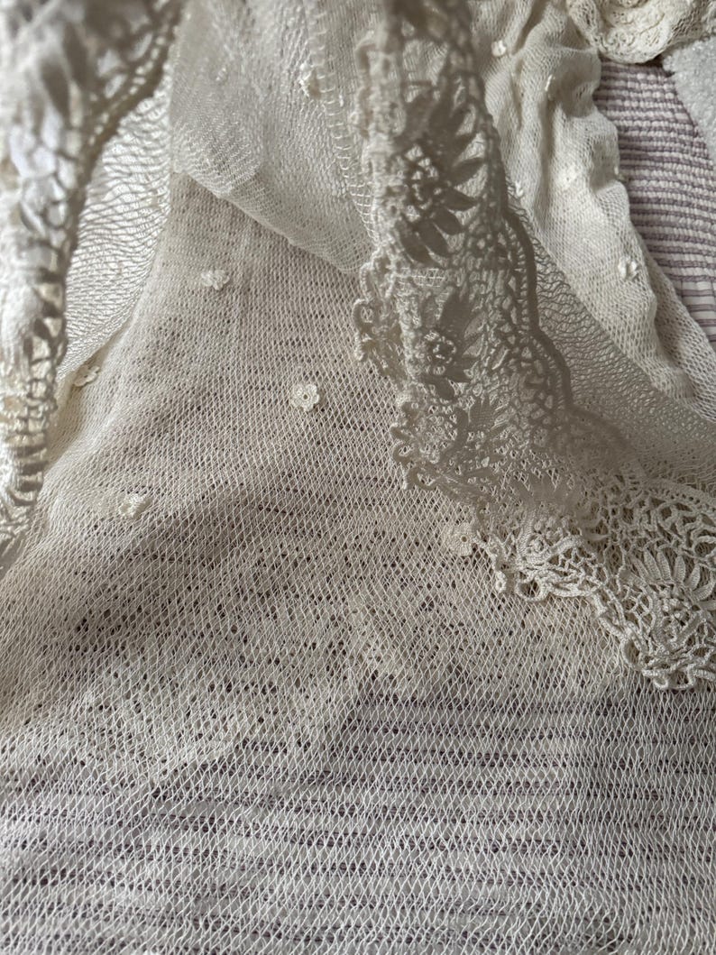 May include: Close-up of a cream-coloured, open-weave fabric with delicate floral lace trim. The fabric has a subtle texture and small, scattered floral embellishments. The lace trim features intricate floral patterns, adding a touch of elegance.