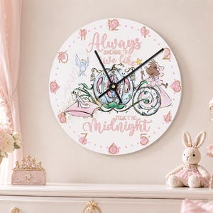 Puede incluir: Un reloj de pared redondo con esfera blanca que presenta una ilustración de una carroza de calabaza azul, un hada madrina y un zapato de cristal sobre un cojín rosa. El texto Always Remember To Live Like There's No Midnight está en rosa.