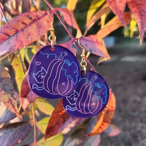 Puede incluir: Pendientes de gato y calabaza de color morado y dorado. Los pendientes son redondos y tienen un gato asomándose por detrás de una calabaza.