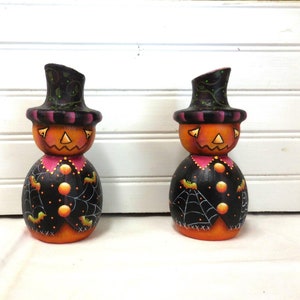 May include: Two Halloween-themed wooden figurines. Each has an orange pumpkin head with a carved face, a black top hat, and a black body decorated with bats and spiderwebs. The figurines are painted with bright colors.