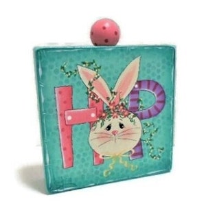 May include: A teal wooden square with the letters "HOP" painted on it. A white bunny with pink ears and a flower crown is in the center of the letters. A pink ball is on top of the square.
