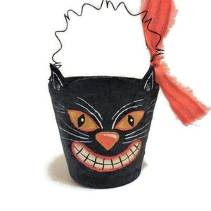 May include: A black cat-faced Halloween bucket with orange eyes, a nose, and a grinning mouth. The bucket has a wire handle and a bright orange fabric tied to it. The design is painted on the bucket, which is set against a white background.