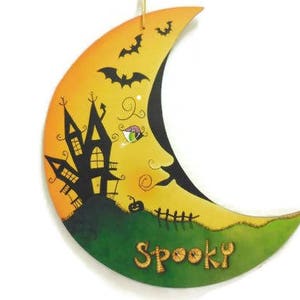 May include: A yellow and green crescent moon with a black silhouette of a house and bats. The moon has a spooky face and the words "Spooky" are written on the bottom.
