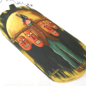 May include: A whimsical, hand-painted Halloween decoration. The wooden plaque features a scarecrow design with orange jack-o'-lantern faces, green pants, and a brown coat. The background is black with a yellow moon and the artist's signature.