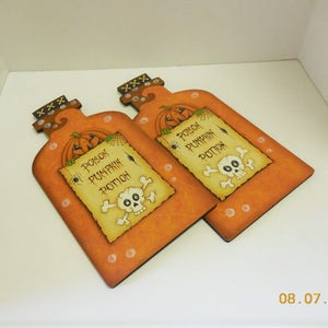 May include: Two wooden Halloween potion bottle decorations. Each bottle is painted orange with a white label that reads "Poison Pumpkin Potion". The labels also feature a skull and crossbones and a spider.