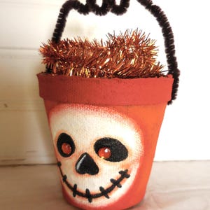 May include: An orange painted terracotta pot decorated with a white skull face featuring black eye sockets, a black nose, and a stitched black mouth. The skull has two orange rhinestone eyes. A brown pipe cleaner handle is attached to the rim.