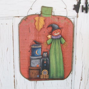 May include: A rustic, pumpkin-shaped wooden sign with a painted Halloween theme. It features a scarecrow holding a star, a black cat, and stacked containers with the words "Happy Fall". The sign is painted in orange, green, black, and blue.