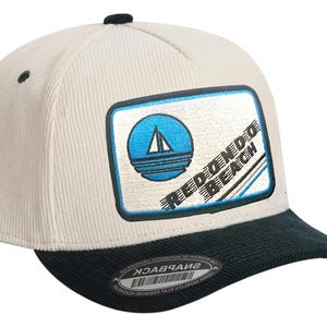 Puede incluir: Gorra de béisbol de pana color crema con visera verde oscuro. La gorra presenta un parche rectangular con un diseño de velero azul y las palabras "REDONDO BEACH" en blanco.