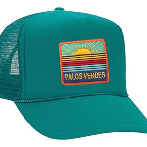May include: Teal trucker hat with a mesh back and a curved brim. The front features a rectangular patch with a sunset design and the text "PALOS VERDES" in yellow.