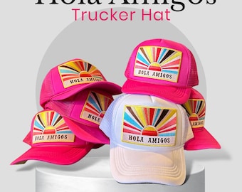May include: Several trucker hats in pink and white. Each hat has a rectangular patch with a colourful sunburst design and the text "HOLA AMIGOS". The hats have a mesh back and a curved peak.