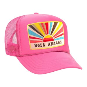 May include: A vibrant pink trucker cap with a mesh back and a curved peak. The front patch displays a colourful sunburst design with the words "HOLA AMIGOS". A bright and cheerful accessory.