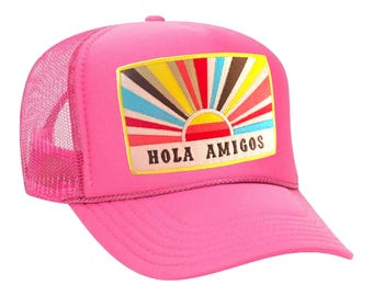 May include: A vibrant pink trucker cap with a mesh back and a curved peak. The front patch displays a colourful sunburst design with the words "HOLA AMIGOS". A bright and cheerful accessory.