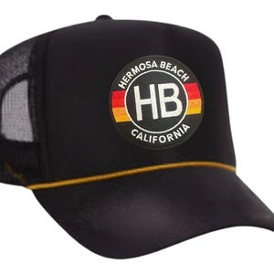 May include: Black trucker hat with a mesh back and a curved peak. The front displays a circular patch with the text "Hermosa Beach California" and "HB" in white, with red, yellow, and orange accents.