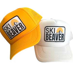 Op de afbeelding: Twee trucker caps met een gele en witte mesh achterkant. Beide caps hebben een zwart-witte patch met de tekst "SKI BEAVER" en een afbeelding van een bergketen en een zon.