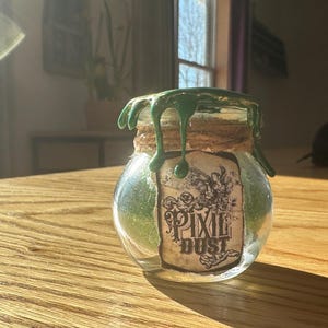 May include: A glass jar with a green wax seal and a label that reads "Pixie Dust". The jar is filled with green glitter and is sitting on a wooden table.