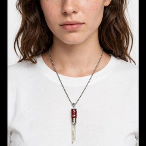 Puede incluir: Un collar de cadena de bolas plateada con un colgante que presenta una base de cristal transparente y puntiaguda. La parte superior del cristal está asegurada por una tapa metálica envuelta en un cordón rojo oscuro, unida a la cadena.