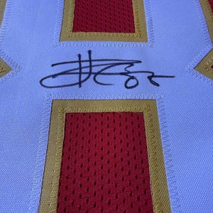 May include: Close-up of a white and red jersey with gold trim. The jersey features a large red square with a mesh pattern, bordered by gold stitching. A black signature is visible on the white fabric.