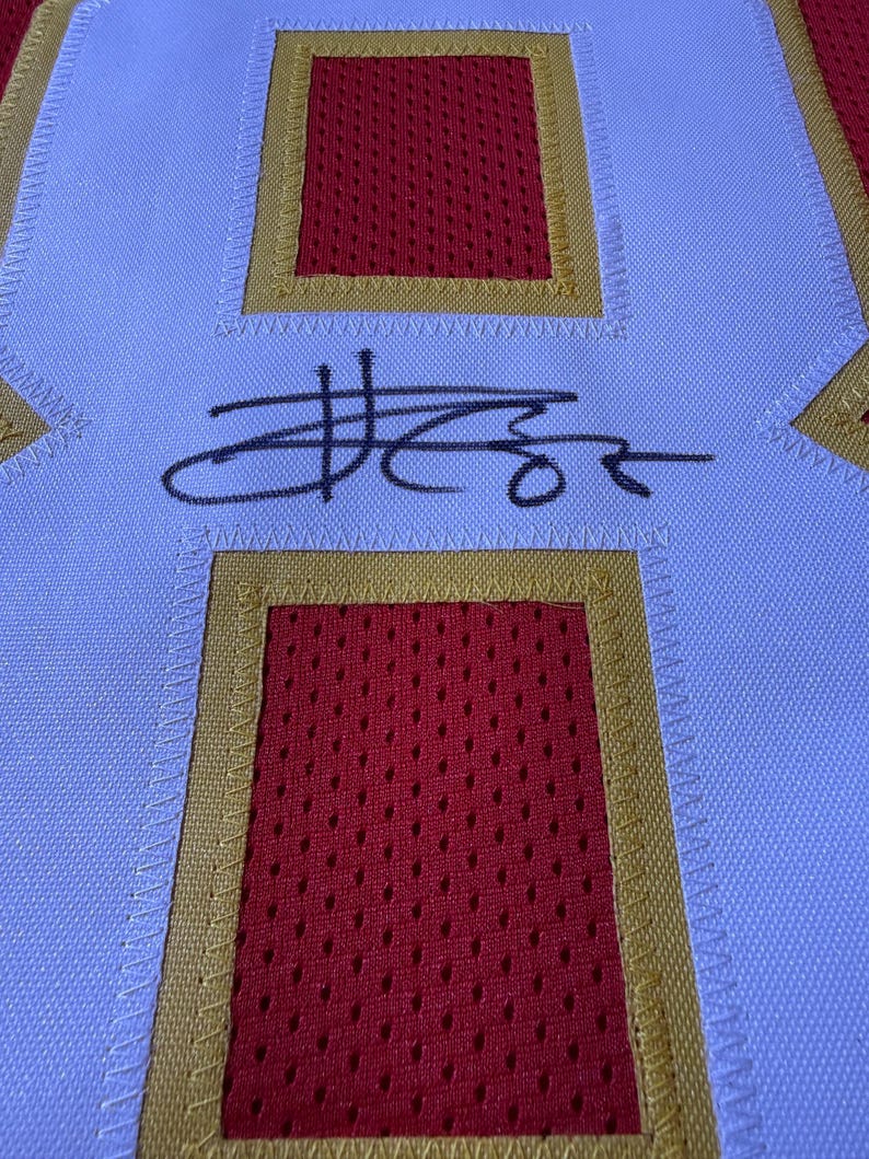 May include: Close-up of a white and red jersey with gold trim. The jersey features a large red square with a mesh pattern, bordered by gold stitching. A black signature is visible on the white fabric.