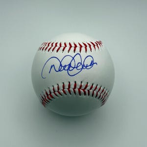 Può includere: Una palla da baseball in pelle bianca con cuciture rosse lungo le giunture. La superficie della palla presenta una firma a inchiostro blu scritta in corsivo al centro, posizionata tra le due file parallele di punti di cucitura rossi.