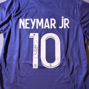Pode incluir: Uma camisa de futebol azul marinho com o nome "NEYMAR JR" e o número "10" em branco. A camisa tem uma assinatura e uma etiqueta Nike. O tecido é brilhante e esportivo.