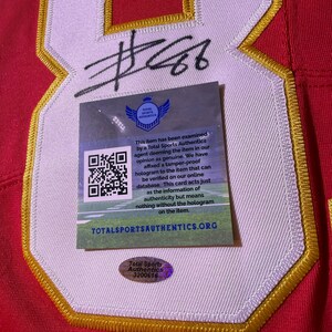 Op de afbeelding: Rood shirt met witte en gouden afwerking, met een groot nummer 8 en een handtekening. Een Total Sports Authentics authenticatiesticker en kaart zijn bevestigd.