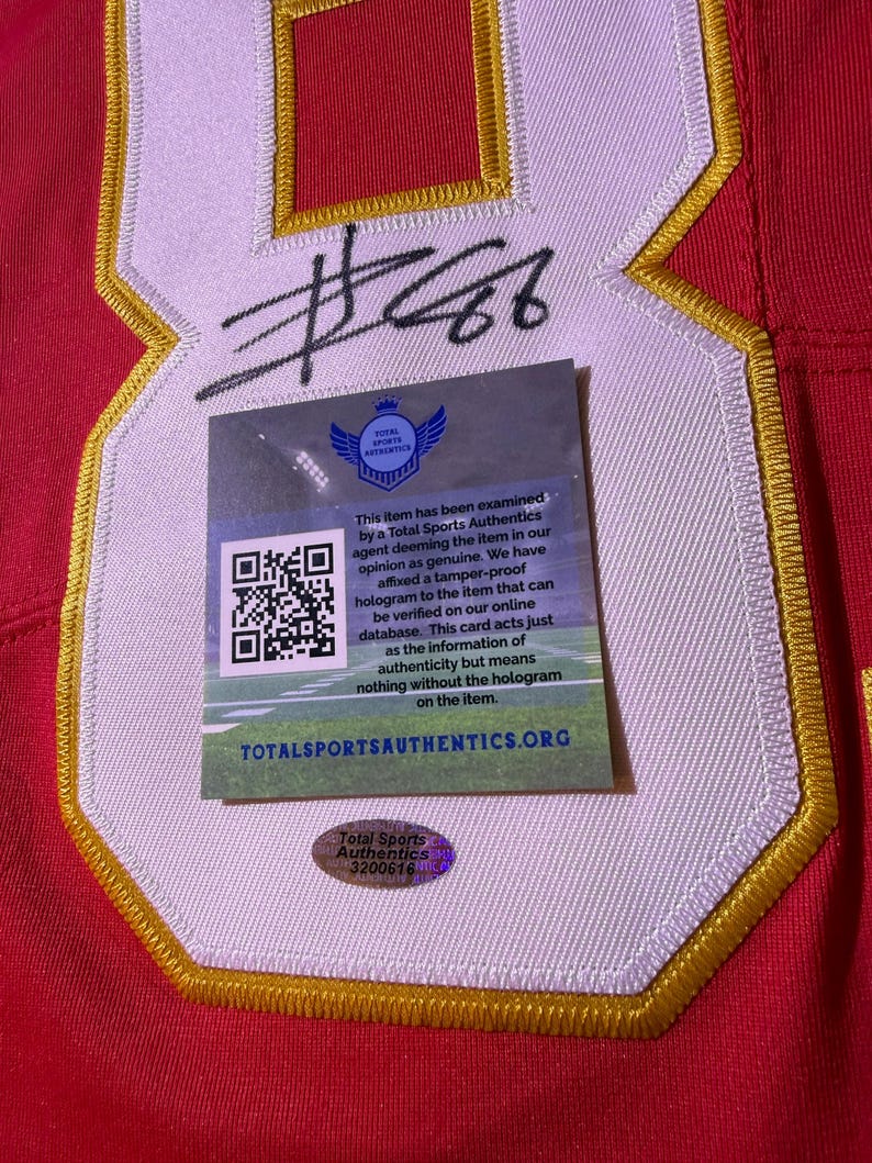 Op de afbeelding: Rood shirt met witte en gouden afwerking, met een groot nummer 8 en een handtekening. Een Total Sports Authentics authenticatiesticker en kaart zijn bevestigd.