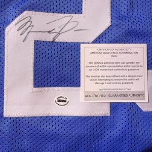 May include: A close-up of a blue mesh jersey with white detailing and a large number. The jersey has an autograph and a certificate of authenticity from American Collectibles Authentication (ACA).