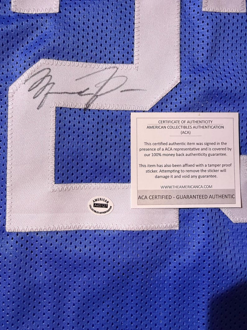 May include: A close-up of a blue mesh jersey with white detailing and a large number. The jersey has an autograph and a certificate of authenticity from American Collectibles Authentication (ACA).
