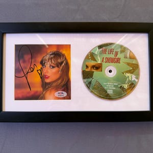 May include: Framed collectible with a signed photograph and a CD. The photograph shows a woman with long hair and a signature. The CD is labelled "THE LIFE OF A SHOWGIRL". The frame is black.