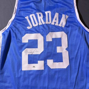 May include: A blue basketball jersey featuring the name "JORDAN" and the number "23" in white. The jersey has white trim around the collar and armholes. A signature is visible below the number.