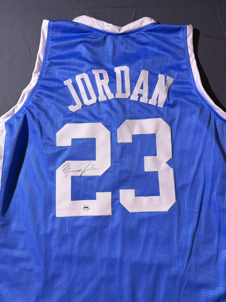 May include: A blue basketball jersey featuring the name "JORDAN" and the number "23" in white. The jersey has white trim around the collar and armholes. A signature is visible below the number.