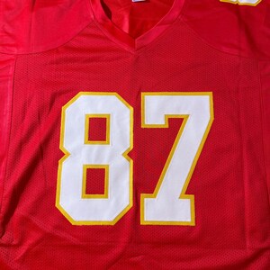 May include: A red football jersey with white and yellow accents. The jersey features the number 87 in large white numerals with yellow outlines on the front. The numbers 7 and 8 are also visible on the shoulders. The jersey has a V-neck.
